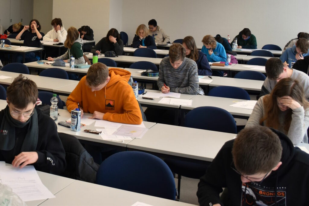 Klausur in der dritten Runde der Mathematik-Olympiade. In einem Klassenraum sitzen Schüler/innen an Tischen und schreiben eine Klausur.